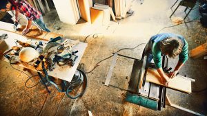 Closeup of unrecognizable adult man slowly pushing wooden plank towards circular saw blade. His coworker on the left is polishing wood with circular sander. Top view, tilt shot.